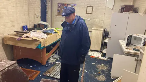 A man wears a blue cap and coat with black gloves and stares solemnly at the ground. He is in a room with debris all over the carpet and mess from vandals breaking in. 