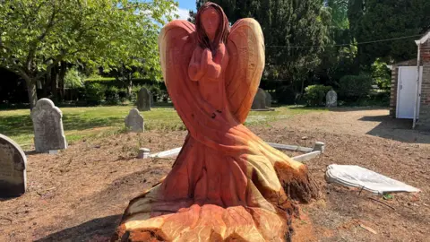 A carved wooden angel made from the stump of a tree. The angel has an orange coloured tan caused by preserving oil. Around the angel are various gravestones with more trees in the background.