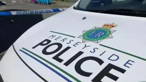BBC Merseyside Police vehicle parked in front of a blue and white police tape cordon. 