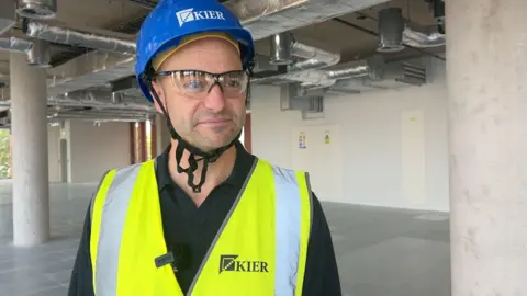 A man in high-vis, a hard hat and protective glasses stands inside an empty building smiling at the camera.