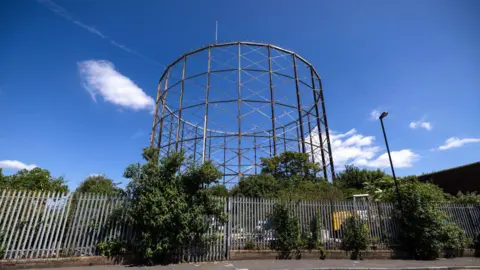 Exterior view of the grade II listed gasholder rising above trees and metal fencing in south London, with its tall iron lattice structure visible against a blue sky.