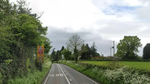 Google A country road with trees and bushes line each side. Street signs and road markings can be seen in the background.