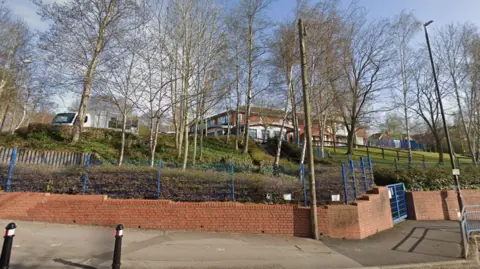 A brick wall with a blue fence on top lines the outside of a school building. There is a small but steep hill with trees on next to the wall with the red-brick school building visible in the background.