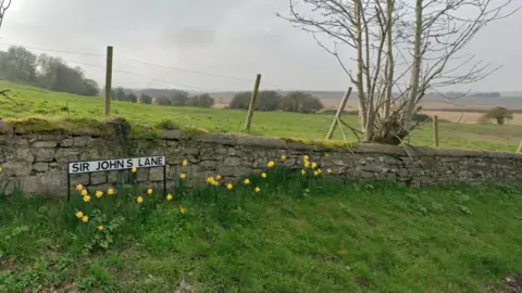 Google A road sign reading Sir John's Lane in Sherburn in Elmet. The sign is in front of a stone wall about one metre high and is surrounded by daffodils. There are open fields in the background. 