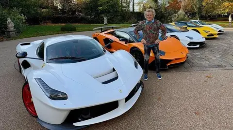 Sir Rod Stewart poses on a large driveway with five sports cars. Three are white, one is yellow and one is orange. Sir Road is standing with his hands on his hips and is wearing ripped jeans, a blue shirt and a patterned jumper