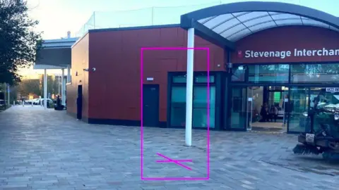 The outside of a bus station, showing a building with a large door and paving outside. There is a pink box, with a cross on the ground, signifying where a statue will be put.