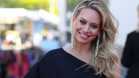 Getty Images Kimberley Wyatt posed in a one shoulder black dress at a red carpet event. her long blonde hair is curled and swept over one shoulder and she is wearing long silver earrings and smiling.