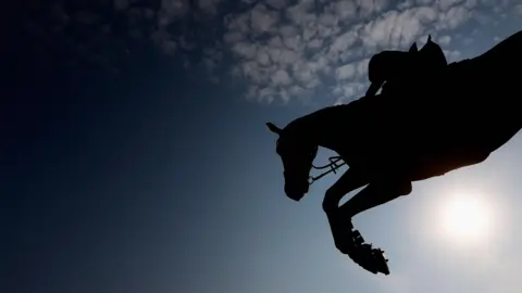 A horse and rider jumping in silhouette against a dark blue sky with high, broken cloud and a hazy white sun.