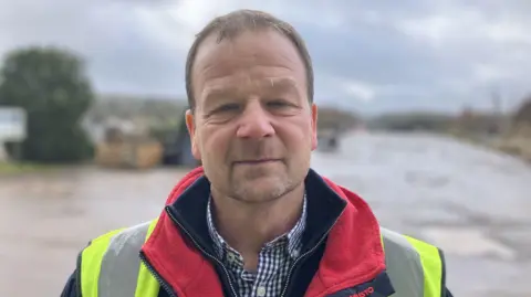 Phil Hall stands in the yard of the company wearing a fleece and high-vis vest. 