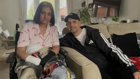 Manjit in a living room, wrapped in blankets with visible medical dressings on their hands and forearms. Kam sits next to her. The background shows household furnishings.