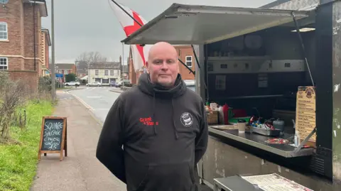 Tony Fisher/BBC A bald man wearing a hoodie with logos standing next to a food van. There is an England flying behind him and a sign on the footpath which reads: "Square sausage, cheese and onion £5". There are homes and cars on the road in the background.