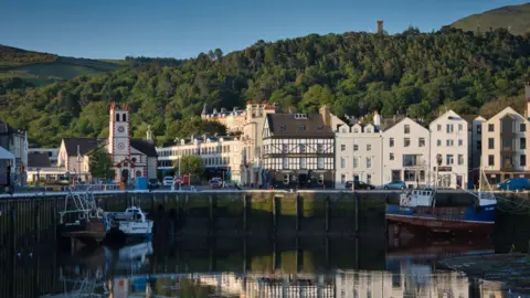 Manx Scenes Ramsey quayside overlooking the harbour, buildings like the quay in front of a leafy hill with the Albert Tower above, on a sunny day.