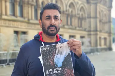 Aisha Iqbal/BBC Shabaz Hussain stands in Centenary Square holding a poster showing a horse, with the words, "He died for your entertainment"