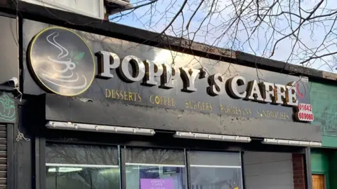 Outside Poppy's Caffe - the name sign above the entrance is in white lettering - there is also a sign saying Desserts Coffee Burgers Parmesans Sandwiches in yellow. There is a logo to the left of this text, of a white cup of tea with steam rising from the top. The background of the sign is black.