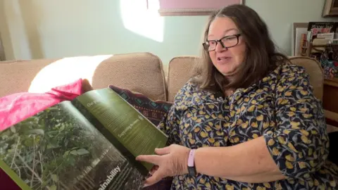 Jane Bertelli, a woman in her mid sixties, sits on the sofa in her home, looking through one of the brochures sent to her by Ethical Forestry Limited.