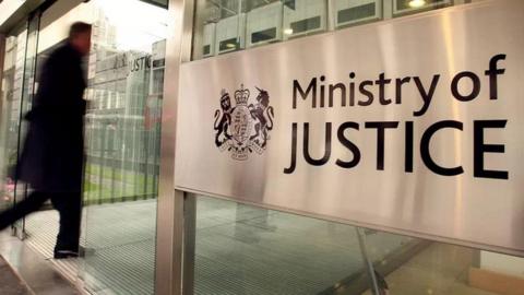 A person in a black coat walking past a sign reading Ministry of Justice