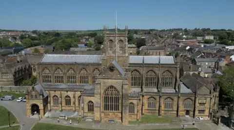 A large stone Abbey with a central tower. There is a grassy area in front of the building that is fronted with large windows. Behind you can see a small town with plenty of leafy trees.