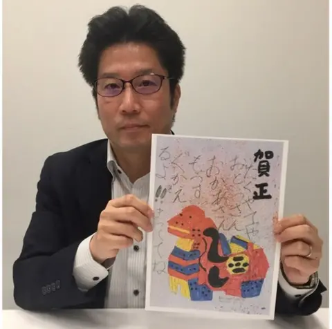 BBC Takuya Yokota, in a black suit, holds up a copy of a postcard featuring a colourful animal