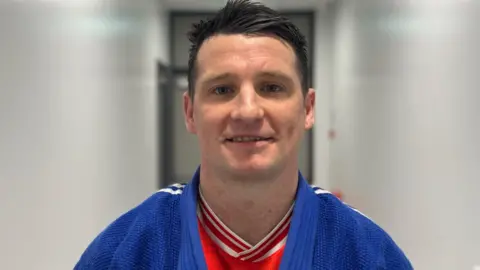 Daniel Grimley has black hair which is swept to the left at the front. He is wearing a blue judo top with a red t-shirt underneath. He is standing in a corridor and is looking at the camera with a small smile on his face. 