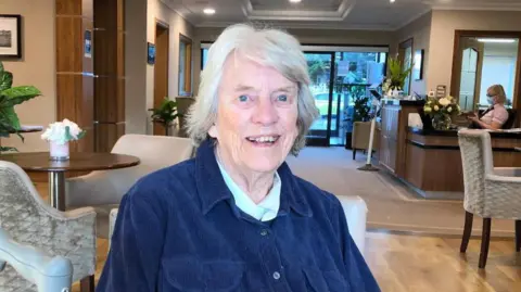 Stella Clarke sitting at a table in what looks like a hotel lobby. She is smiling into the camera.