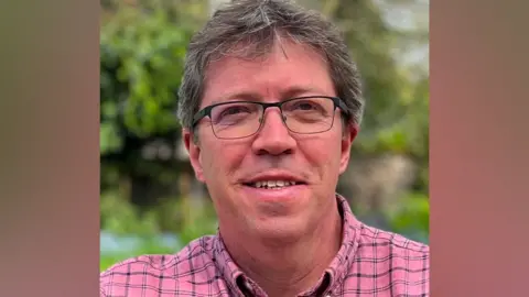 Liberal Democrats A man wearing glasses with a pink stripped shirt and grey-speckled dark hair smiles towards the camera, with the background greenery blurred