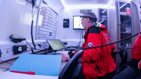 Tim Wood in the Cleveland Mountain Rescue control vehicle. He is wearing a red waterproof jacket and a head torch, and is looking at a map on a computer screen.