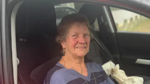 George King/BBC Nina Ceretti sitting in the driver's seat of her car. She is wearing a striped jumper with gold chain. She is smiling while looking into the camera.