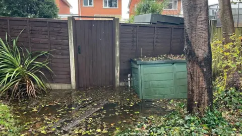 Photo of a dark wooden fence with a gate in the middle, about 6-feet high. At the bottom of the fence is a large puddle of water with leaves and branches floating on the surface. To the right of gate is a large green plastic garden container, and beyond it is a two-storey red brick house with white window frames. 