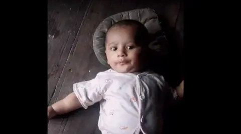 Handout Fayaz's son looks at the camera. The baby is lying on a wooden surface with a pillow behind his head, wearing a baby grow. 