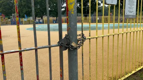 A padlock and chain are across metal gates with a drained paddling pool in the background.