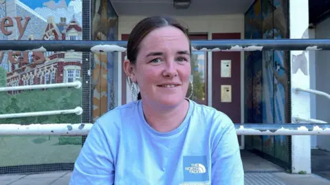 A woman wearing a blue top is looking at the camera and smiling. There are black railings behind her and she has silver hoop earrings.