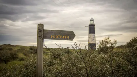 A wooden signpost has yellow text stating 'footpath' on it. A black and white striped lighthouse and the sea can be seen in the background on an overcast day.