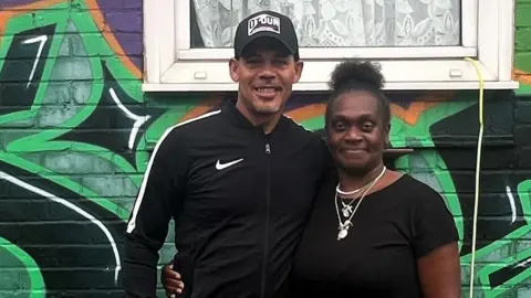 Vincent Nembhard, left, stands next to Sandra Nembhard. They both are wearing black and have one arm wrapped around each other. They are standing in front of a mural on their garden wall which says the word 'stretch' in black and green graffiti font on the bottom.