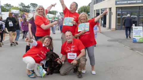 A Gorman Photography Five adults wearing red shirts with the words "ask me" on them strike a pose in the middle of a walking route at the Bath and West Show with a building and other people in the background.