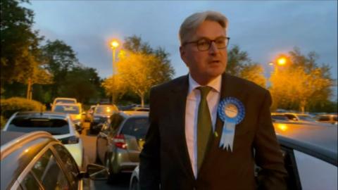 Daniel Kawczynski standing in a car park next to several cars. He wears a dark suit over a white shirt with a green tie and a blue rosette pinned to his suit. He has white, grey hair and wears glasses