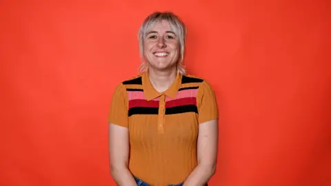 Maisie Adam wearing a yellow striped top with pink and red on it. She has blonde short hair and is standing in front of a red background. 