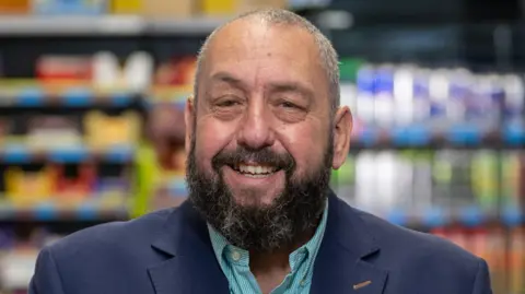 A man smiling at the camera. He is wearing a blue blazer and turquoise shirt, and has a dark beard and greying hair. Supermarket fridge shelving is behind him out of focus