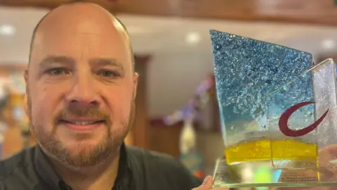 A man who is bald with a beard wearing a black shirt and holding up an award which is made of glass that is half blue and half yellow