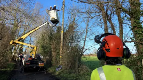 A van with a platform is elevating two electricians, dressed in Hi-Viz and Helmets. They are working on a brown electricity poll. They are approximately 20 feet in the air. Either side of the poll are tree's. A man in a red helmet and Yellow Hi-Viz stands beside the camera with his back to it. 