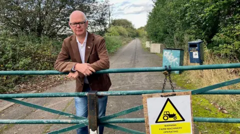 Councillor Stuart Jeffery from Maidstone Borough Council. Stuart is leaning against a green gate. He is wearing jeans and a brown jacket. 