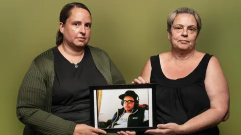 ALEX POWELL The picture shows the upper bodies of two women, Carole and Linda, sitting down in front of a green wall. They are holding a photo of Chris Carpenter in a black frame. This is an upper body picture of Chris, wearing glasses, a hat and headphones.