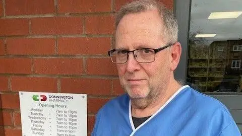 Nigel Dugmore A man with black framed glasses and grey hair with a blue medical top on stood beside a brick wall with Donnington Pharmacy and opening hours written on a sign