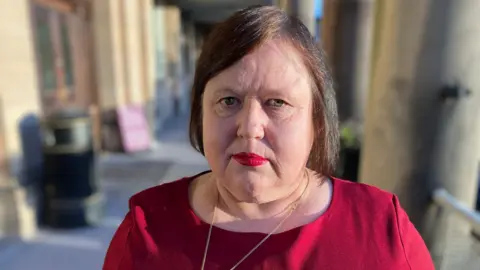 Joan Dixon the leader of the Labour Group on Derbyshire County Council standing infront of county hall in Matlock in a red top 