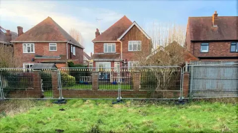 Mark Palmer A view from inside the field towards a row of three red brick detached houses.  Heras fencing has been installed along the perimeter line of their back gardens, running parallel to a low garden wall. 