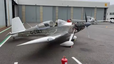 A silver aircraft is grounded outside a warehouse storage area. The plane is silver and has a small cabin with a glass roof.