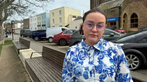 BBC/Jadzia Samuel Shelli is sitting on a park bench, with cars parked up behind