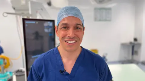 A surgeon wearing blue surgical scrubs and a theatre cap stands in an operating room, with medical equipment and monitors visible in the background.
