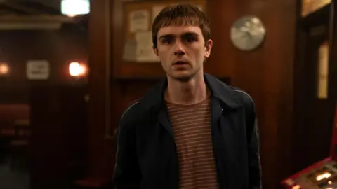 BBC/Mam Tor Productions/Anne Binckebanck Mitchell Robertson in character as Young Niall. He looks fearful and nervous, stood alone in a bar. He is wearing a navy tracksuit top with a red and white striped t-shirt underneath. A gambling machine is visible behind him, as is the wooden-cladded interior of the bar.