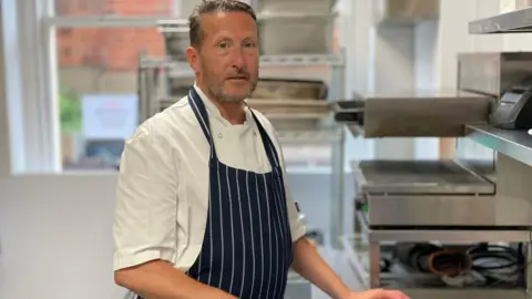A chef wearing a white shirt and a striped black and white apron. He is standing near a stove. 