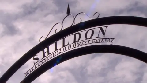 A large metal arch with the words "Shildon 200, Millennium East Gateway" with blue sky and clouds behind.